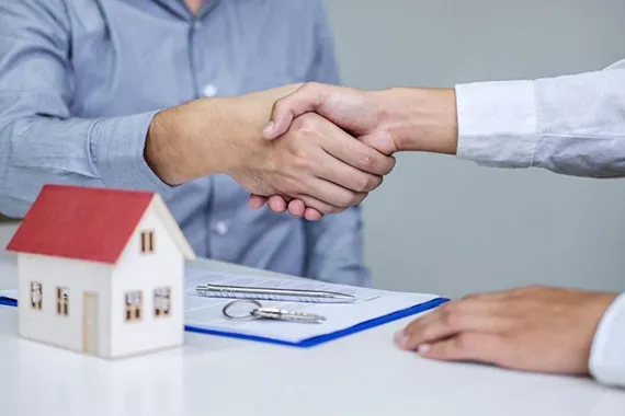 Two professionals shaking hands over a real estate contract with a small red-roofed house model, illustrating the benefits of landlord loans.